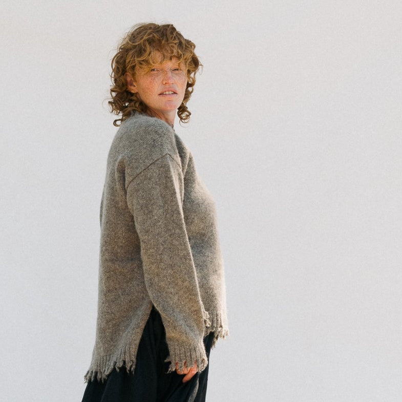 Person wearing a gray sweater and black pants standing on a wooden block against a white background