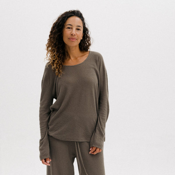 A woman modeling a matching lounge set consisting of a long-sleeve crewneck top and wide-leg cropped pants in a taupe-brown color, paired with light gray slip-on shoes, against a plain white studio background.