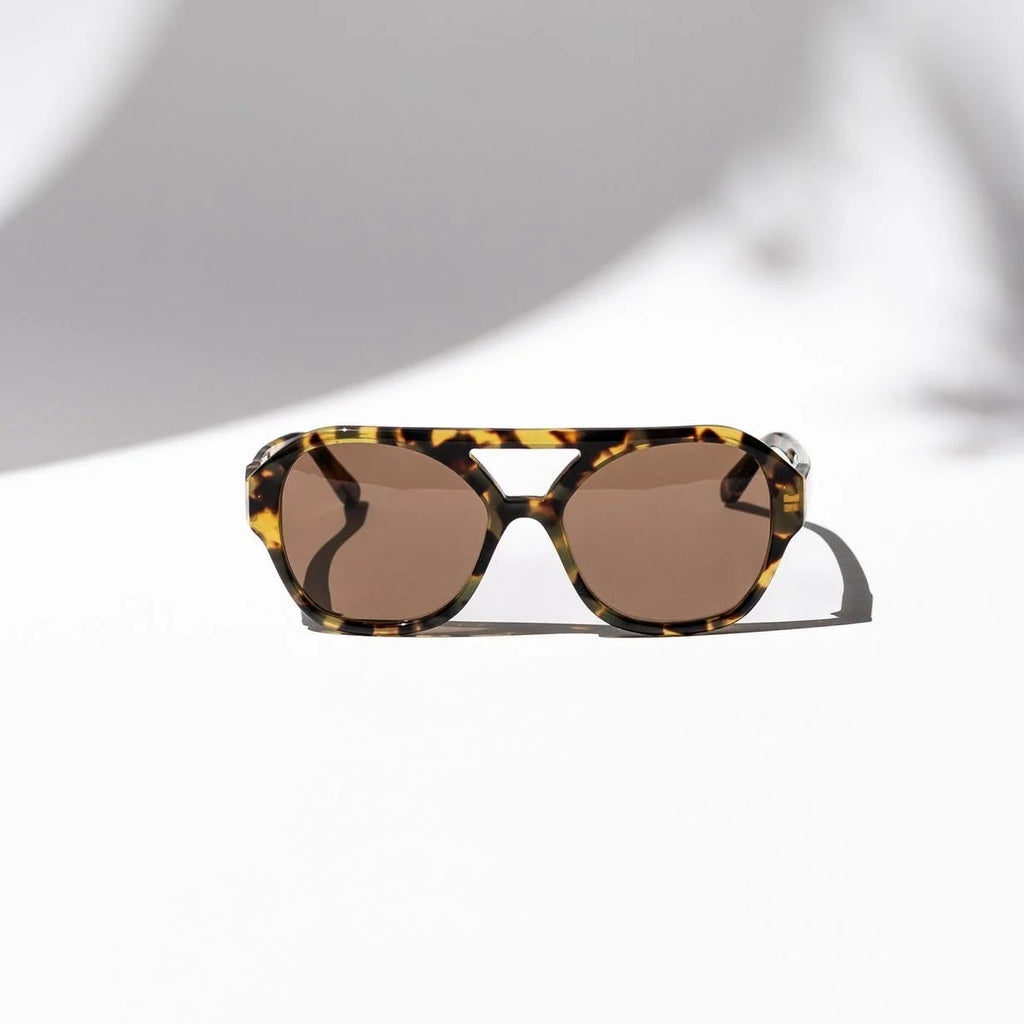 A pair of sunglasses with a tortoiseshell patterned frame and brown lenses, resting on a white surface with artistic shadows.