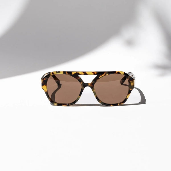 A pair of sunglasses with a tortoiseshell patterned frame and brown lenses, resting on a white surface with artistic shadows.