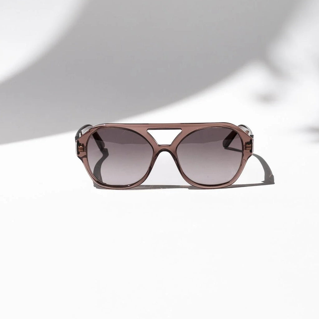 A pair of cat-eye sunglasses with a translucent rose gold frame and gradient gray lenses, resting on a white surface with artistic shadows.