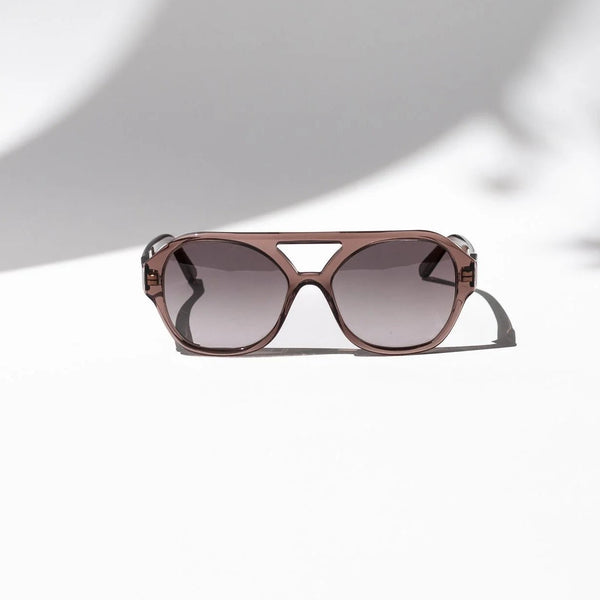 A pair of cat-eye sunglasses with a translucent rose gold frame and gradient gray lenses, resting on a white surface with artistic shadows.