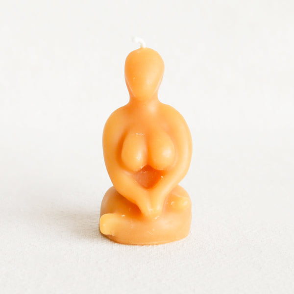 A beige-colored candle in the shape of a sitting woman figurine, with a smooth and slightly glossy finish.